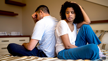 A man and woman sit on a bed, both displaying expressions of disappointment and concern